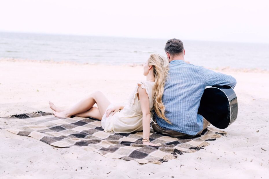 Core values list | Back view of a man and woman on the beach looking out to sea. They lay on a blanket leaning against each other. She wears a white dress and he wears a denim shirt and holds a guitar.