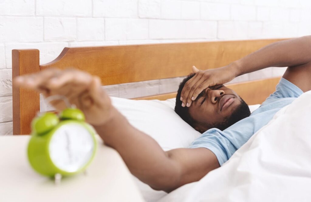 Lucid dreaming: hit the snooze button | A man lays in bed with his hand over his eyes and his hand on the top of a green alarm clock.