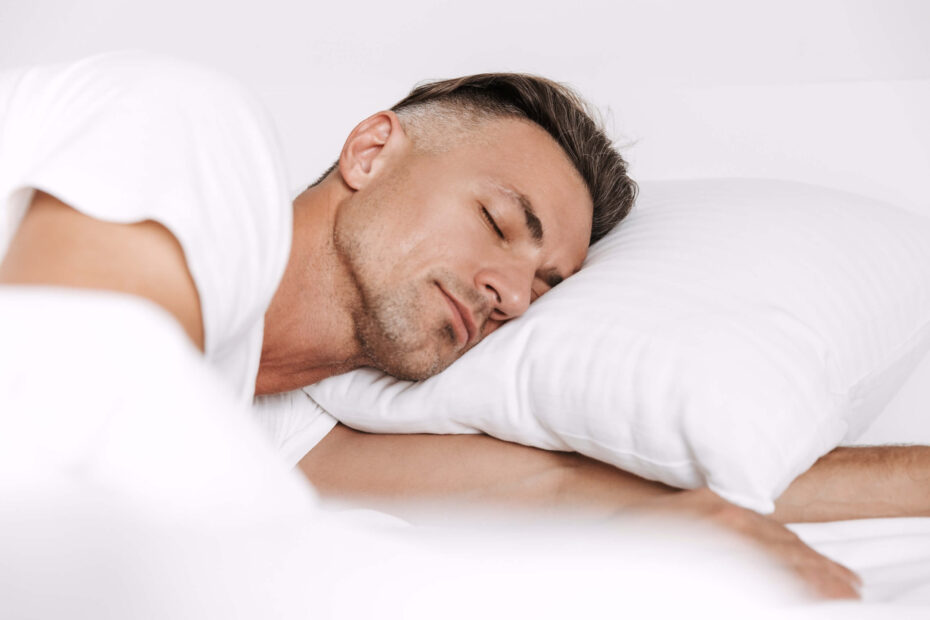 Psychology of dreams | A middle-aged man with greying hair sleeps in a bed with white bedding. He's wearing a white t-shirt.