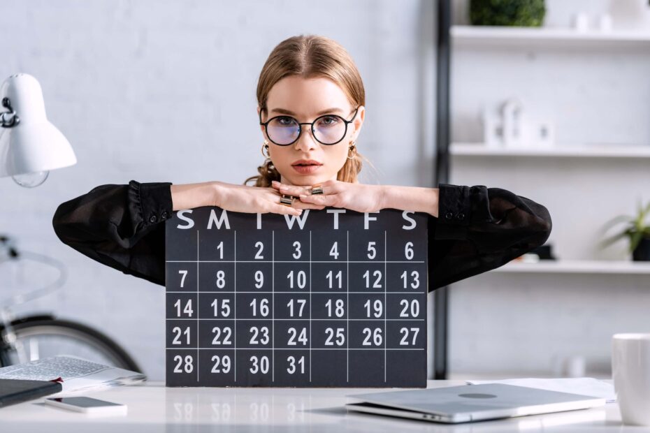 30-day challenges | A woman wearing glasses and black cardigan sits at a desk with her arms resting on top of a chalk board calendar.