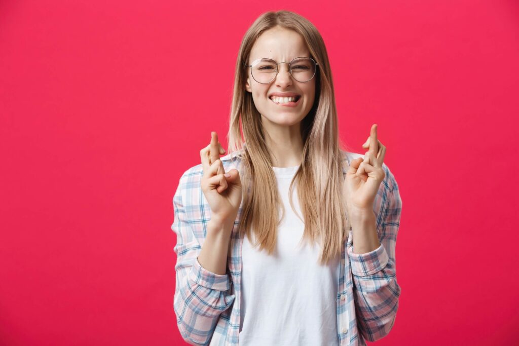 The psychology of superstitions and symbolism | A blonde lady crossing her fingers against a pink background.